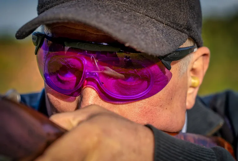 An experienced clay pigeon shooter wearing X Sight Sport shooting glasses with prescription RX inserts. The glasses feature a purple lens, providing enhanced contrast and target visibility. The shooter is seen mounting his gun, focusing on the target ahead. With X Sight Sport's premium eyewear, shooters can enjoy improved vision and protection during their shooting activities.