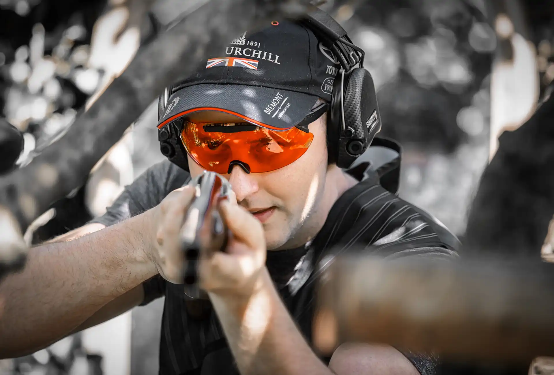 A male clay shooter, wearing ear protection and X Sight Sport shooting glasses with a vibrant orange lens, gazes down the barrel of his shotgun aimed towards the camera. The bold orange lens serves as the focal point of the image, enhancing contrast and visibility. The backdrop of trees creates a dynamic sporting atmosphere, reflecting the challenging nature of the sport. This captivating shot captures the intensity and determination of the shooter, ready to take on the clay shooting discipline.