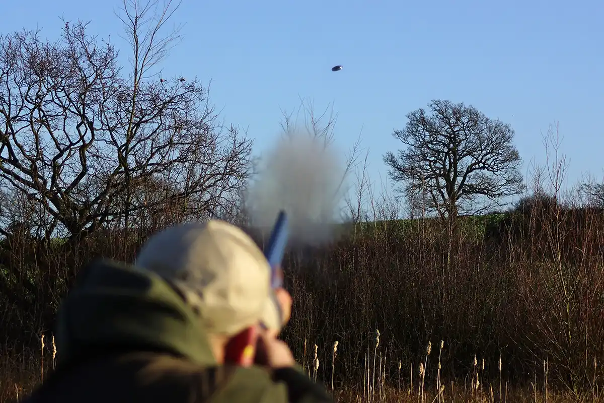Image displays a clay shooter from behind, at a sporting shoot shooting at a black clay target being thrown away into a blue sky. The secne is clearly wintery and the light looks quite dim.