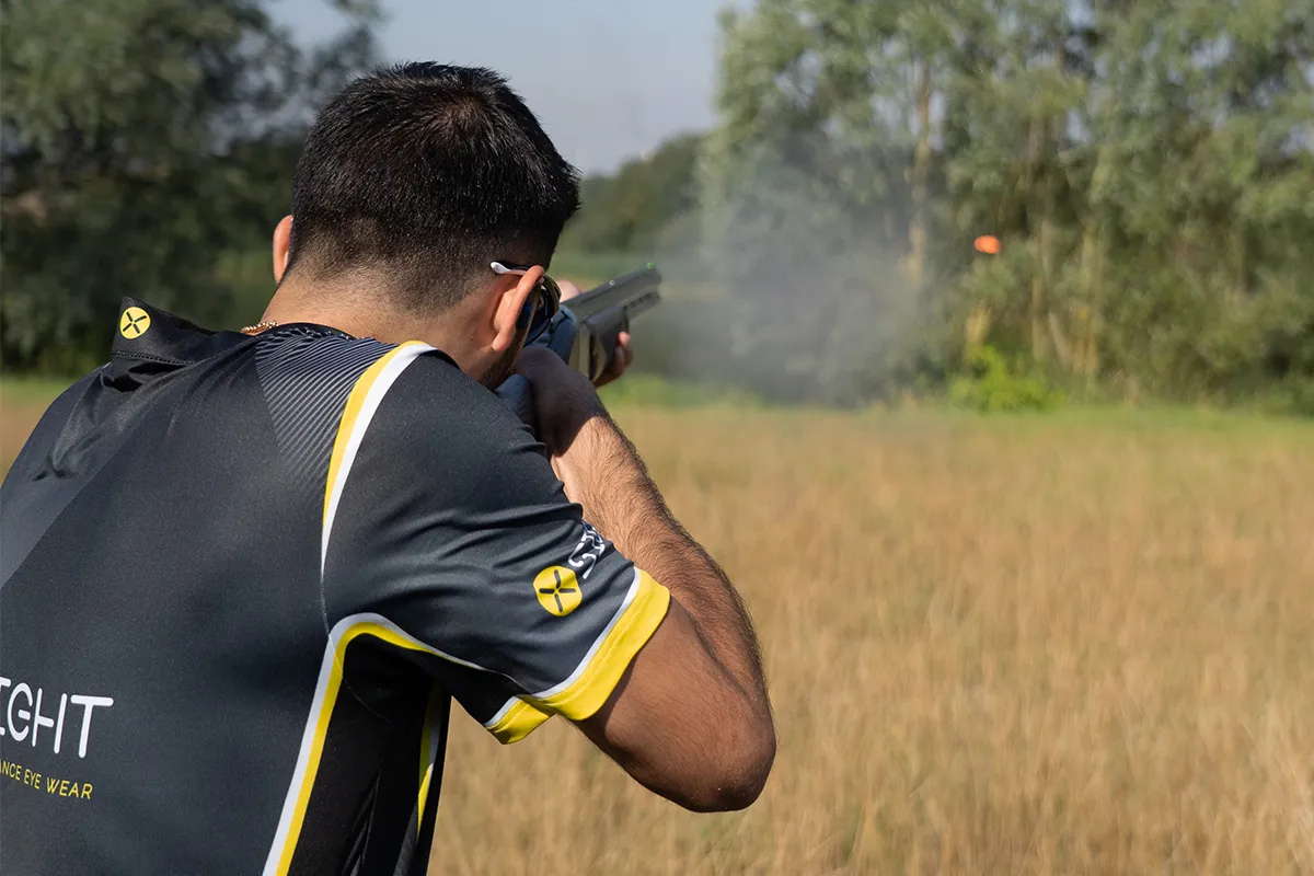 Image displays a clay shooter from behind, at a clay shooting ground shooting the discipline of English Sporting where he is shooting at an orange clay target in the distance being thrown from across a backdrop of greenery and trees.