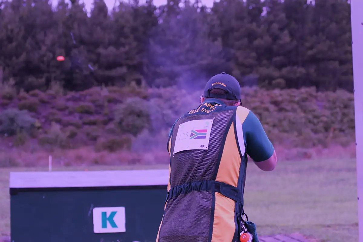 The image shows how the following scene would look when viewed through the Deep Purple lens from X Sight Sport shooting glasses. The scene displays a trap shooter from behind, at a trap shooting ground aiming and shooting at an orange clay target being thrown from the trap house against a green background of treeline and hillside.