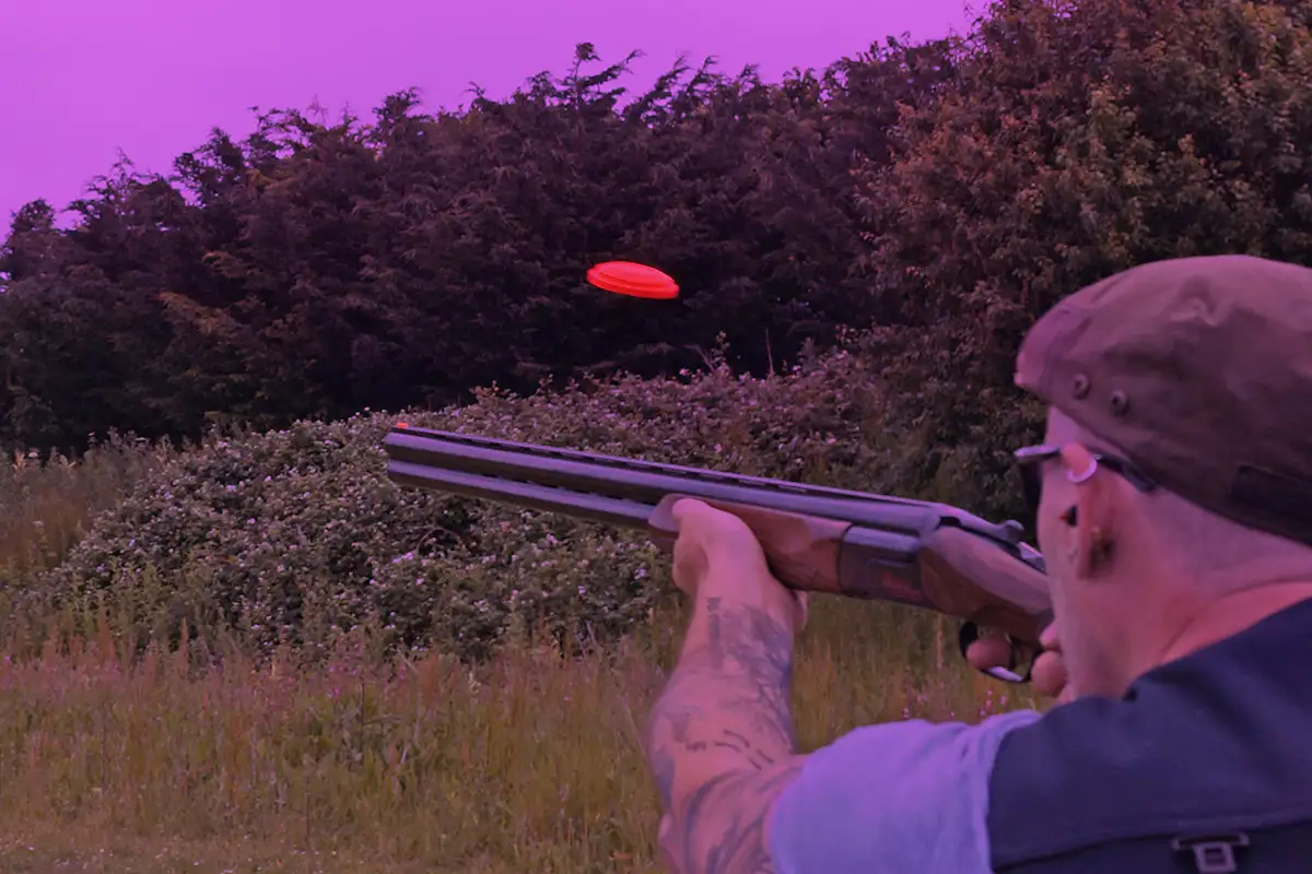 The image shows how the following scene would look when viewed through the Electric Purple lens from X Sight Sport shooting glasses. The scene displays a clay shooter from behind, aiming and about to shoot at an orange clay target against green shrubbery and treeline. He is pointing his gun in the direction of where the clay is going.