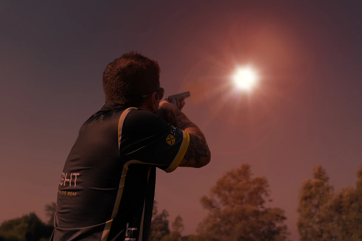 The image shows how the following scene would look when viewed through the HD Brown lens from X Sight Sport shooting glasses. Image displays a clay shooter from behind wearing an X Sight shooter shirt, aiming and shooting at a clay target in the sky that is directly passing the sun. It was too bright to see the clay before but now with the HD Brown lens you can see it clearly. The trajectory of the shot is visible. The sky is a deep shade of purple brown and there are trees at the bottom in the background.