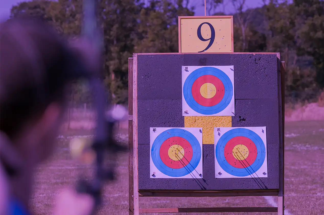 Image captured through the Deep purple lens color of X Sight Sport shooting archery glasses. The photo features a compound archery target with three full-color 50m target faces during an archery competition. From behind the compound archer, the target is in focus, while the archer is blurred out. This image vividly depicts how the scene and target appear through the lens, highlighting the enhanced clarity and color enhancement provided by the lens color. The photo is part of an image comparison slider, which shows the original photo and a realistic visual representation of the view offered by lens colour.