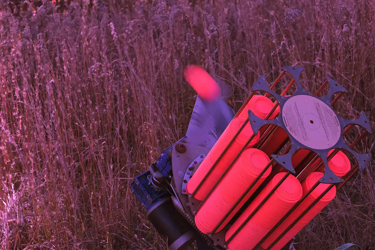The image shows how the following scene would look when viewed through the light pink lens from X Sight Sport shooting glasses. The scene shows a clay pigeon trap angled towards the sky. It is full of orange claze coloured clay targets. One is being thrown. it is blurred showing the speed and motion of the clay. The background behind the trap is brown long tall grass.